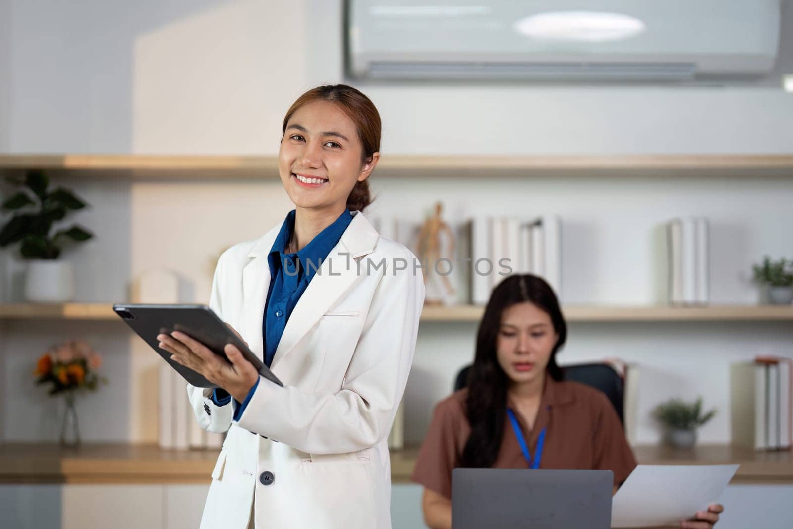 A woman confidently presenting ideas while her colleague focuses on work, showcasing leadership and teamwork in a modern office.