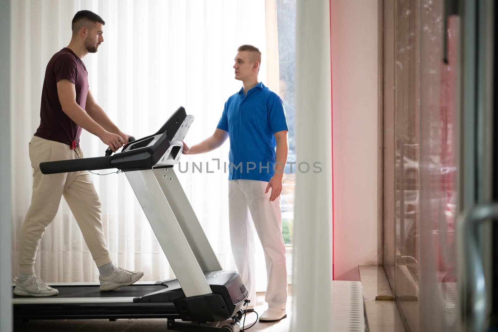 Man undergoing physical therapy with trainer assistance on a treadmill indoors.