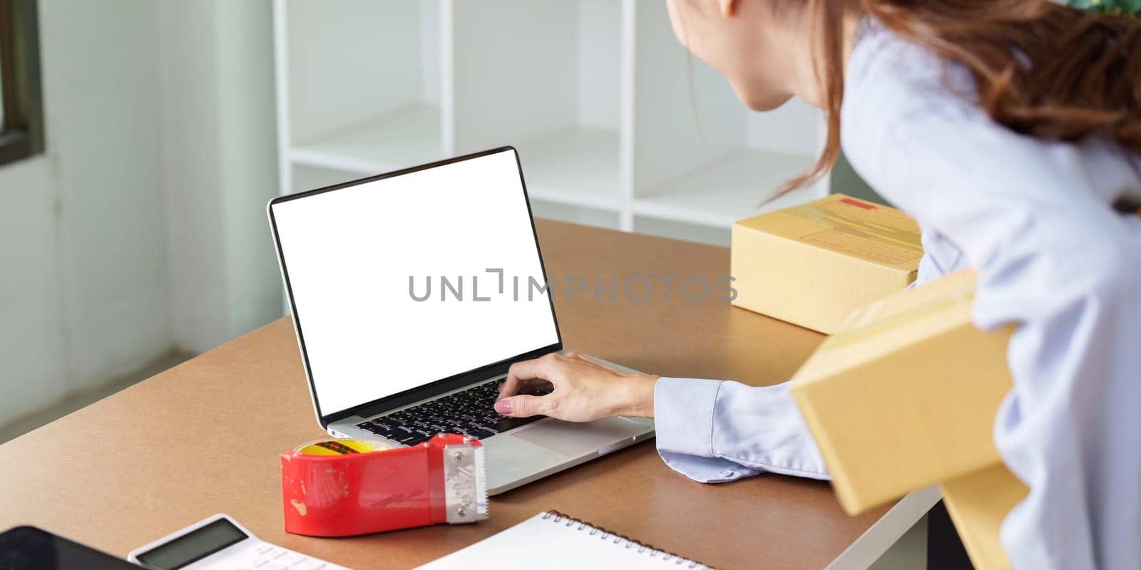 A businesswoman engages with her laptop while managing packages, showcasing the dynamics of small business operations.