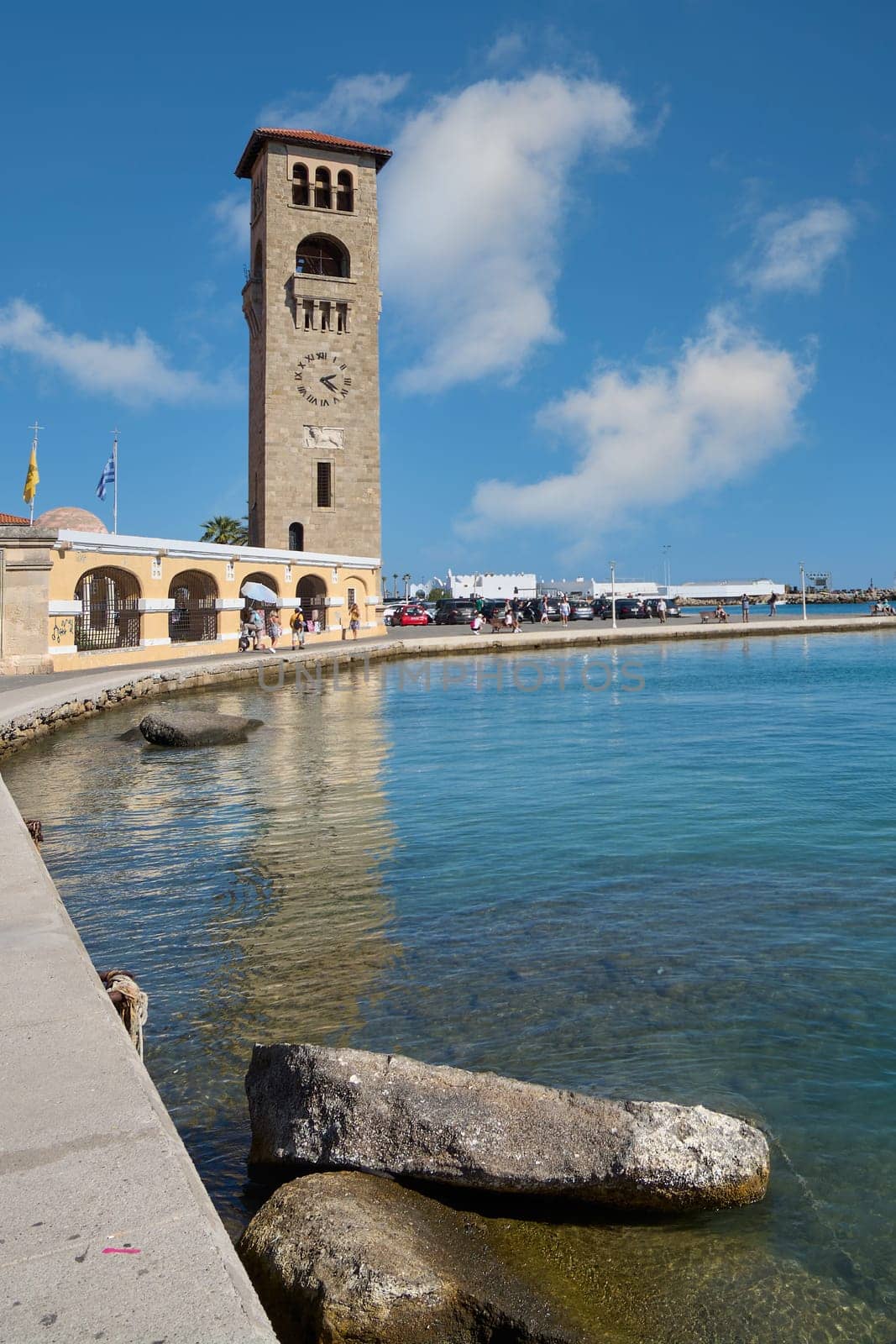 Rodas.Greece - june 04, 2025: The imposing clock tower of Rhodes by Mandraki Harbour, a prominent landmark reflecting on the calm azure waters.