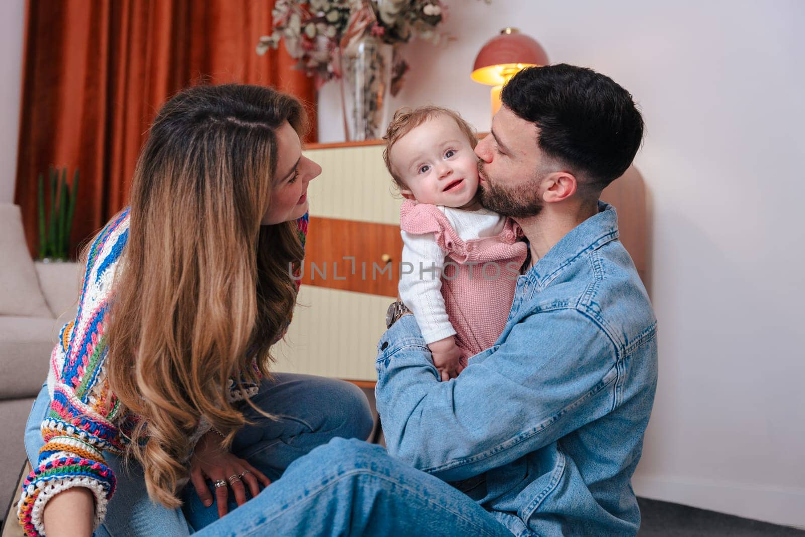 Touching family moment with father holding and kissing his baby girl as mother looks on. Captured in a cozy indoor space, ideal for themes like love, childhood, parenting, and emotional bonds.