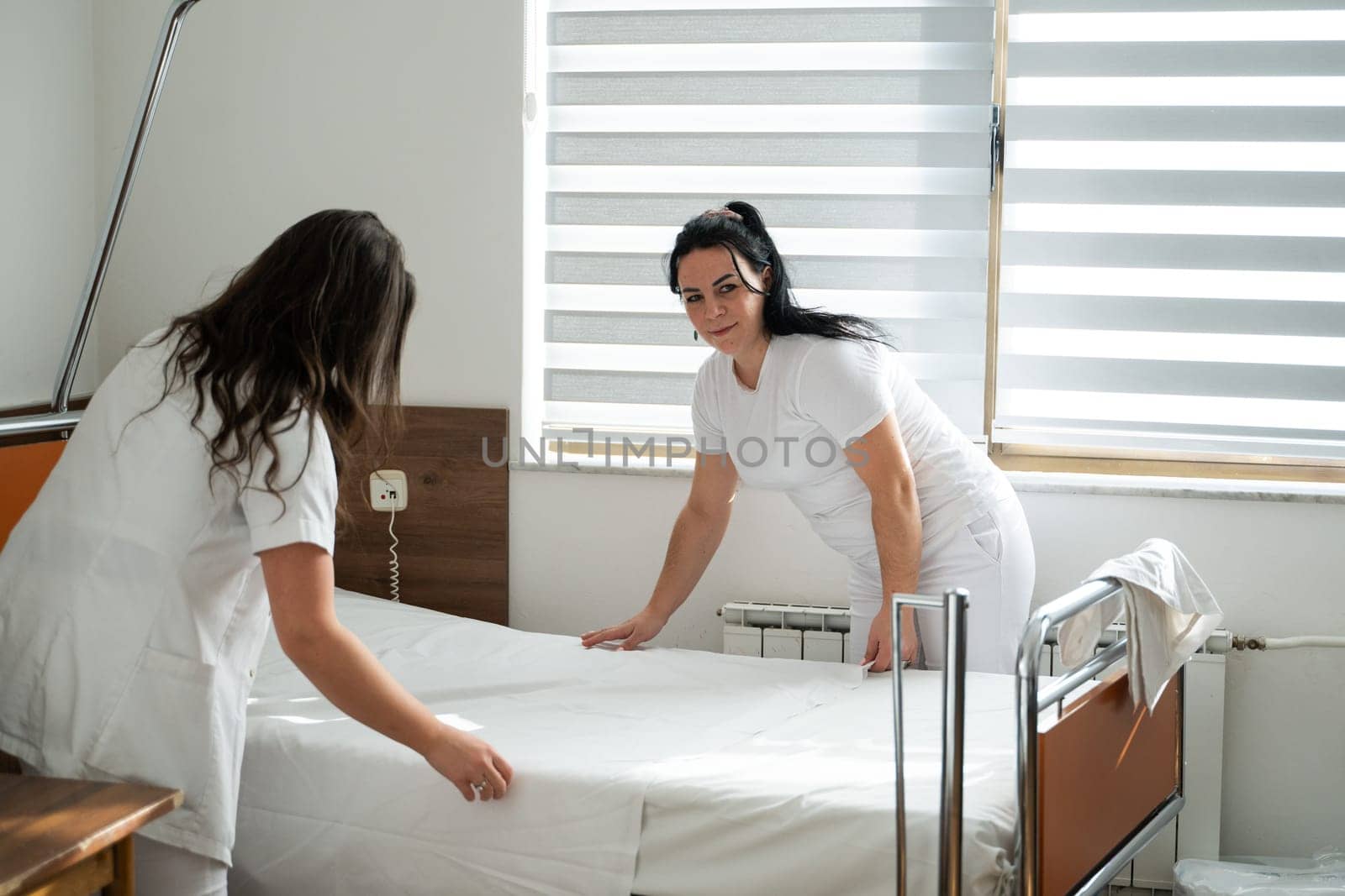 Two nurses in uniforms cleaning and organizing a hospital room by changing the bed linens and ensuring a hygienic environment for patients