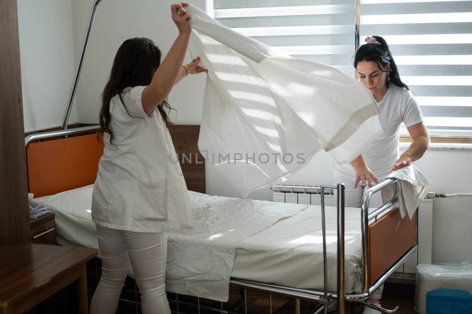 Two nurses in uniforms cleaning and organizing a hospital room by changing the bed linens and ensuring a hygienic environment for patients