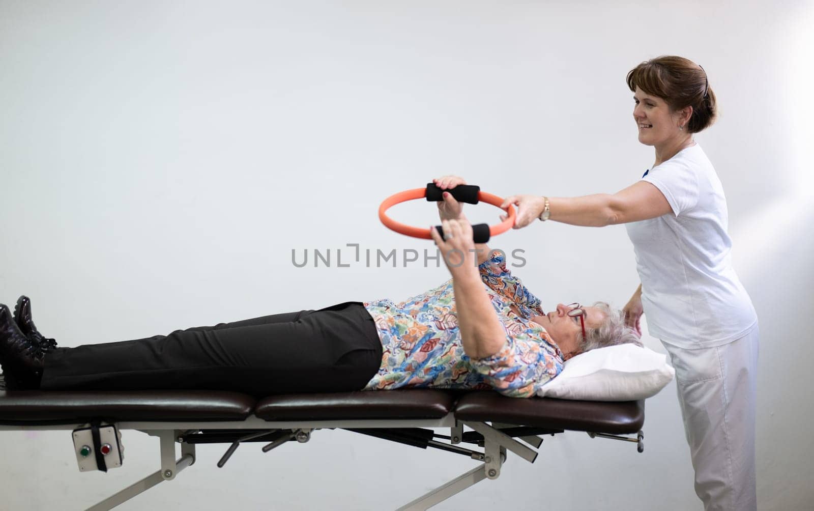 Elderly woman undergoing physiotherapy under the supervision of a female therapist. The patient is lying on a treatment table while performing an arm exercise with an orange pilates ring.