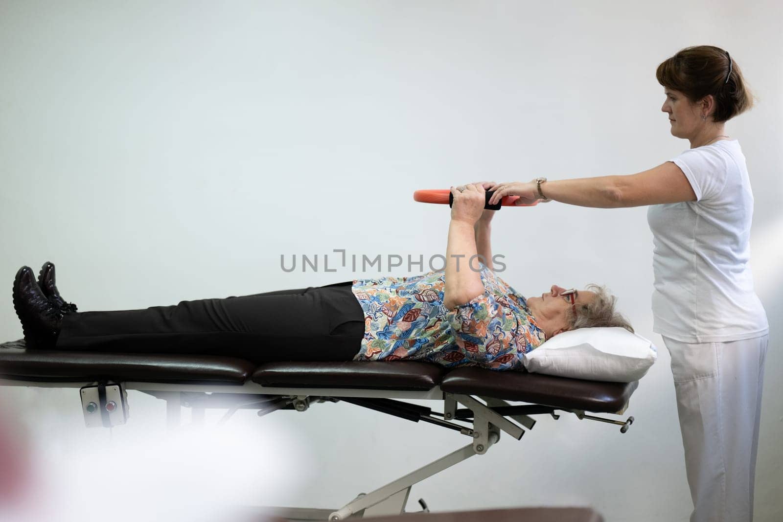 Elderly woman undergoing physiotherapy under the supervision of a female therapist. The patient is lying on a treatment table while performing an arm exercise with an orange pilates ring.
