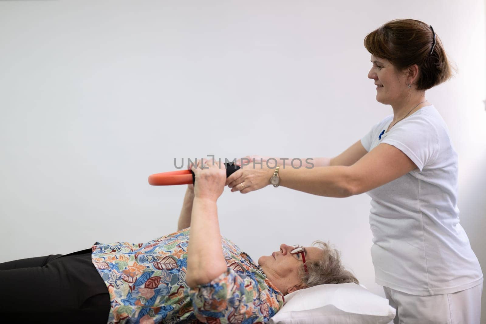 Elderly woman undergoing physiotherapy under the supervision of a female therapist. The patient is lying on a treatment table while performing an arm exercise with an orange pilates ring.