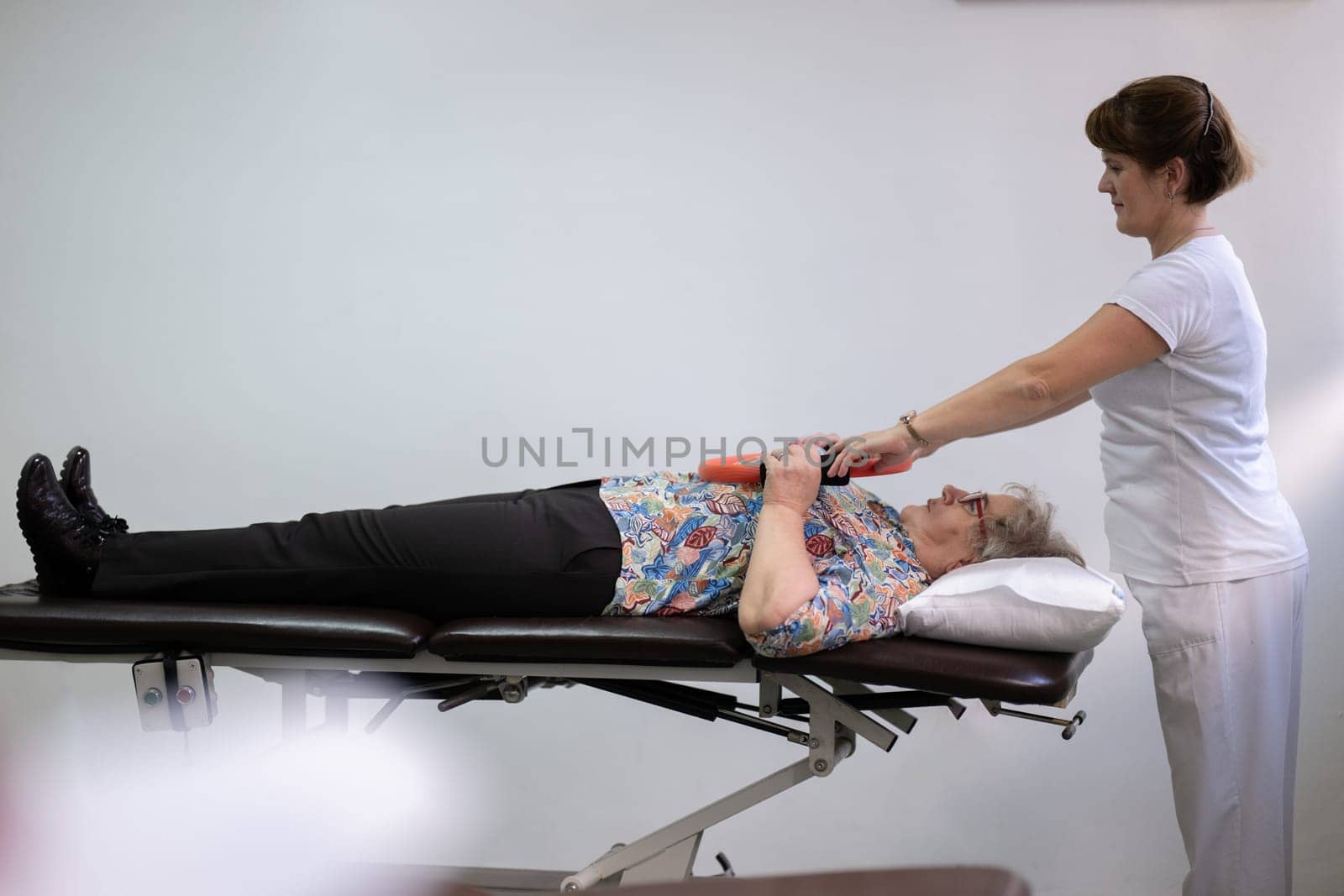 Elderly woman undergoing physiotherapy under the supervision of a female therapist. The patient is lying on a treatment table while performing an arm exercise with an orange pilates ring.