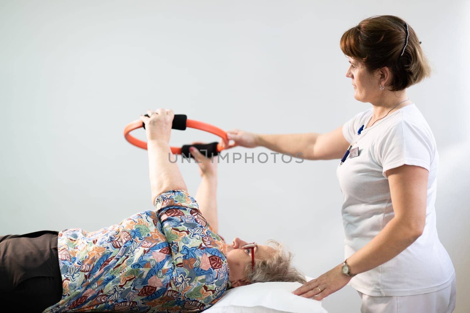 Elderly woman undergoing physiotherapy under the supervision of a female therapist. The patient is lying on a treatment table while performing an arm exercise with an orange pilates ring.