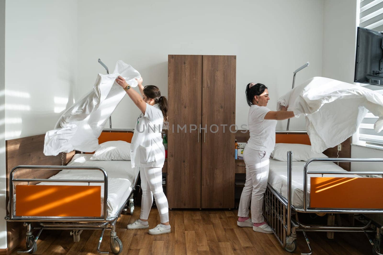 Two nurses in uniforms cleaning and organizing a hospital room by changing the bed linens and ensuring a hygienic environment for patients