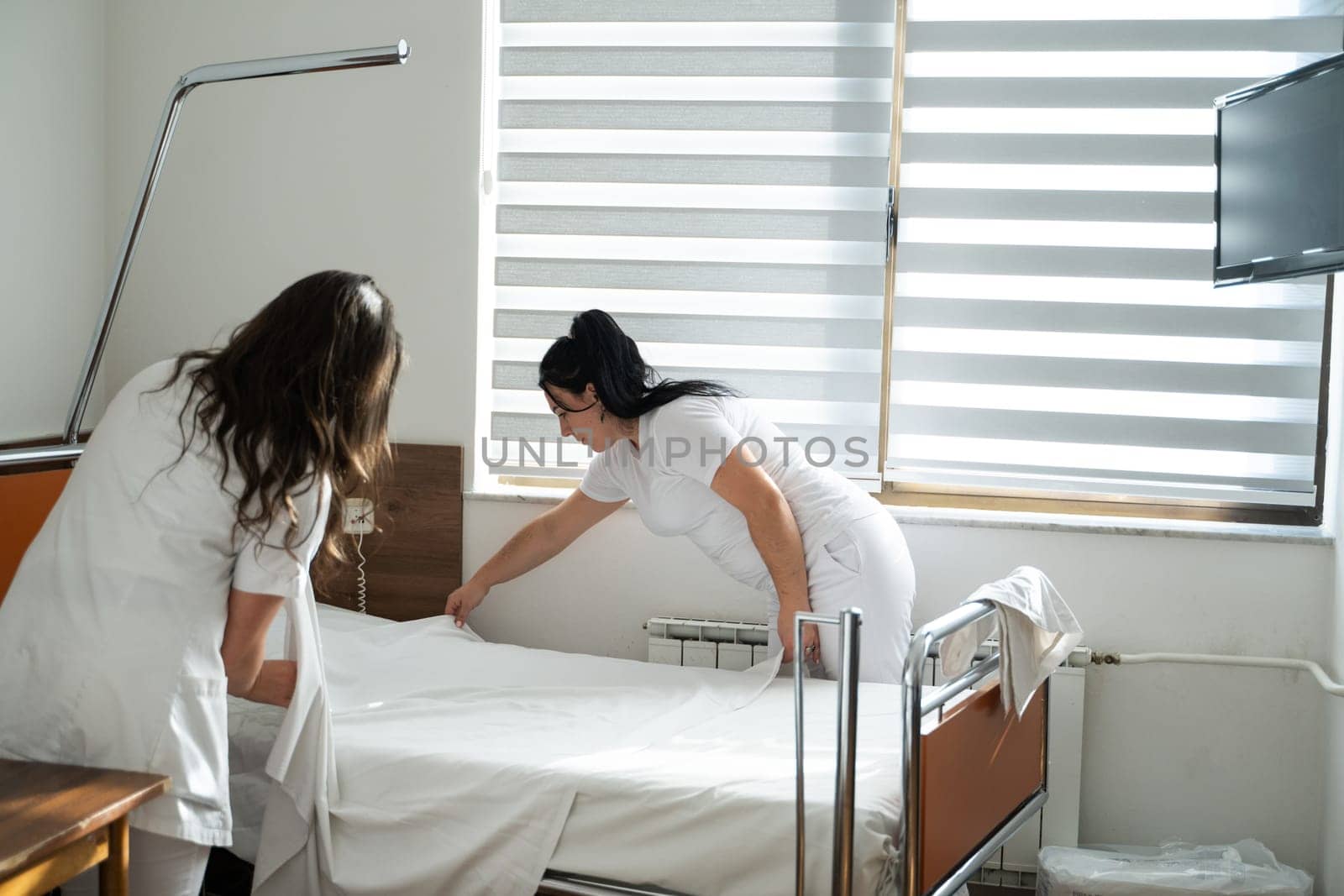 Two nurses in uniforms cleaning and organizing a hospital room by changing the bed linens and ensuring a hygienic environment for patients