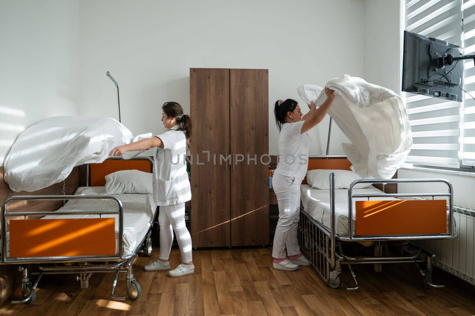 Two nurses in uniforms cleaning and organizing a hospital room by changing the bed linens and ensuring a hygienic environment for patients