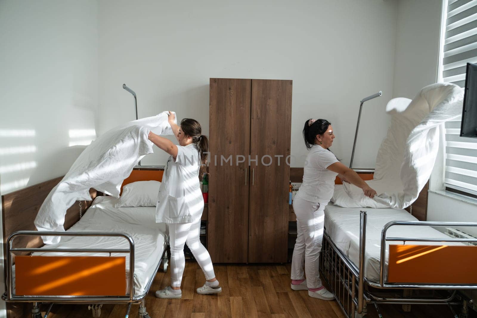 Two nurses in uniforms cleaning and organizing a hospital room by changing the bed linens and ensuring a hygienic environment for patients
