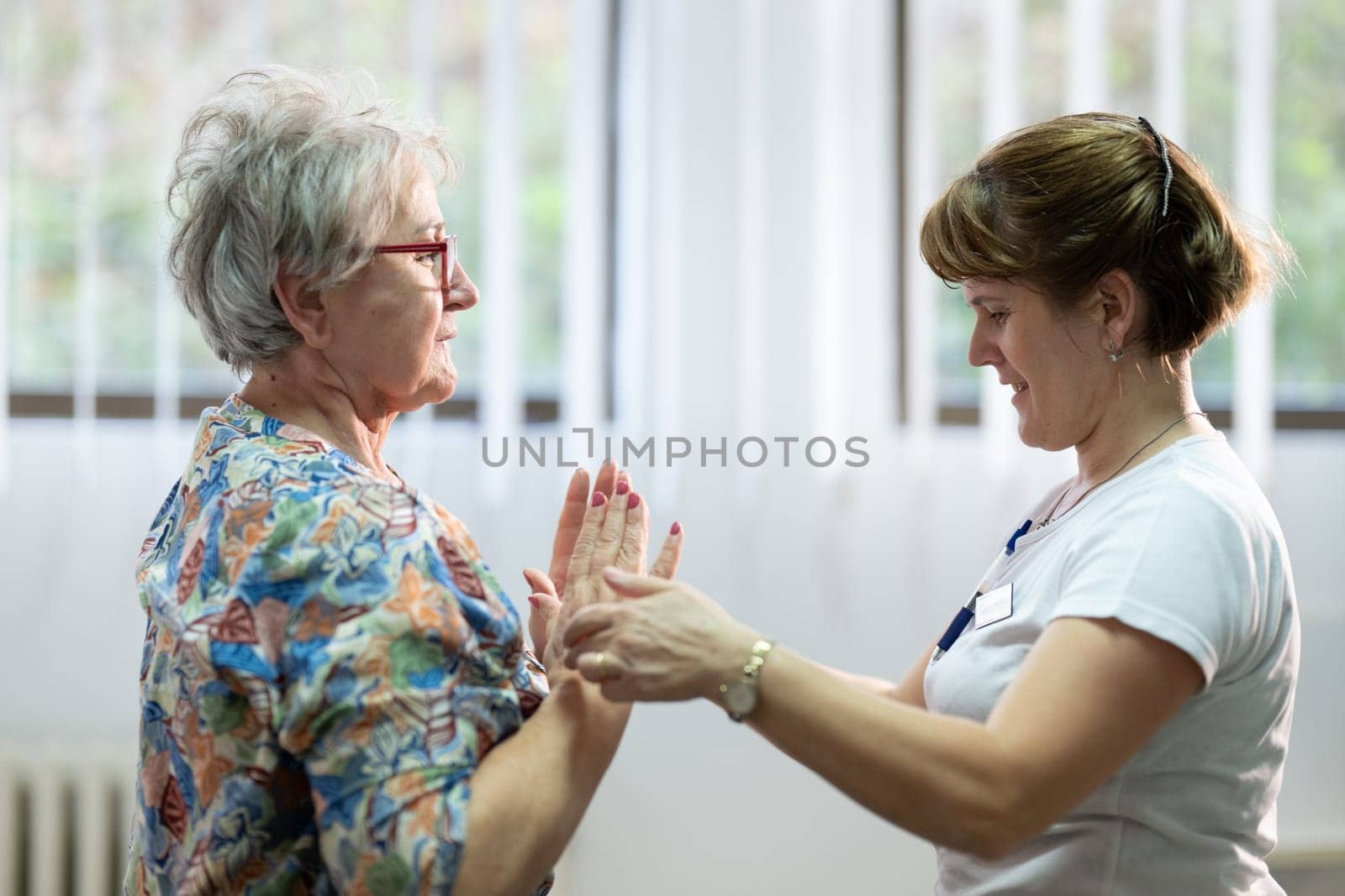 Senior woman doing physical exercises with the help of a smiling therapist. Concept of elderly rehabilitation and movement recovery.