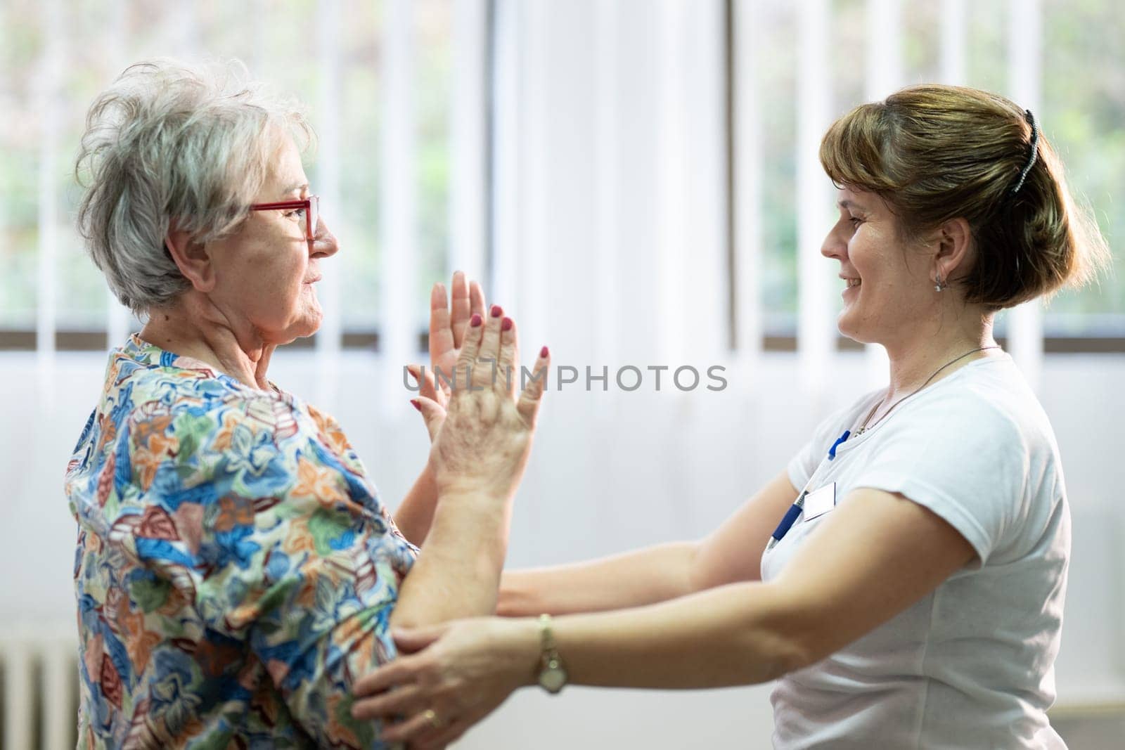 Senior woman doing physical exercises with the help of a smiling therapist. Concept of elderly rehabilitation and movement recovery.