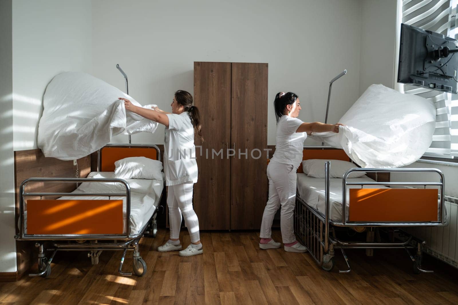 Two nurses in uniforms cleaning and organizing a hospital room by changing the bed linens and ensuring a hygienic environment for patients