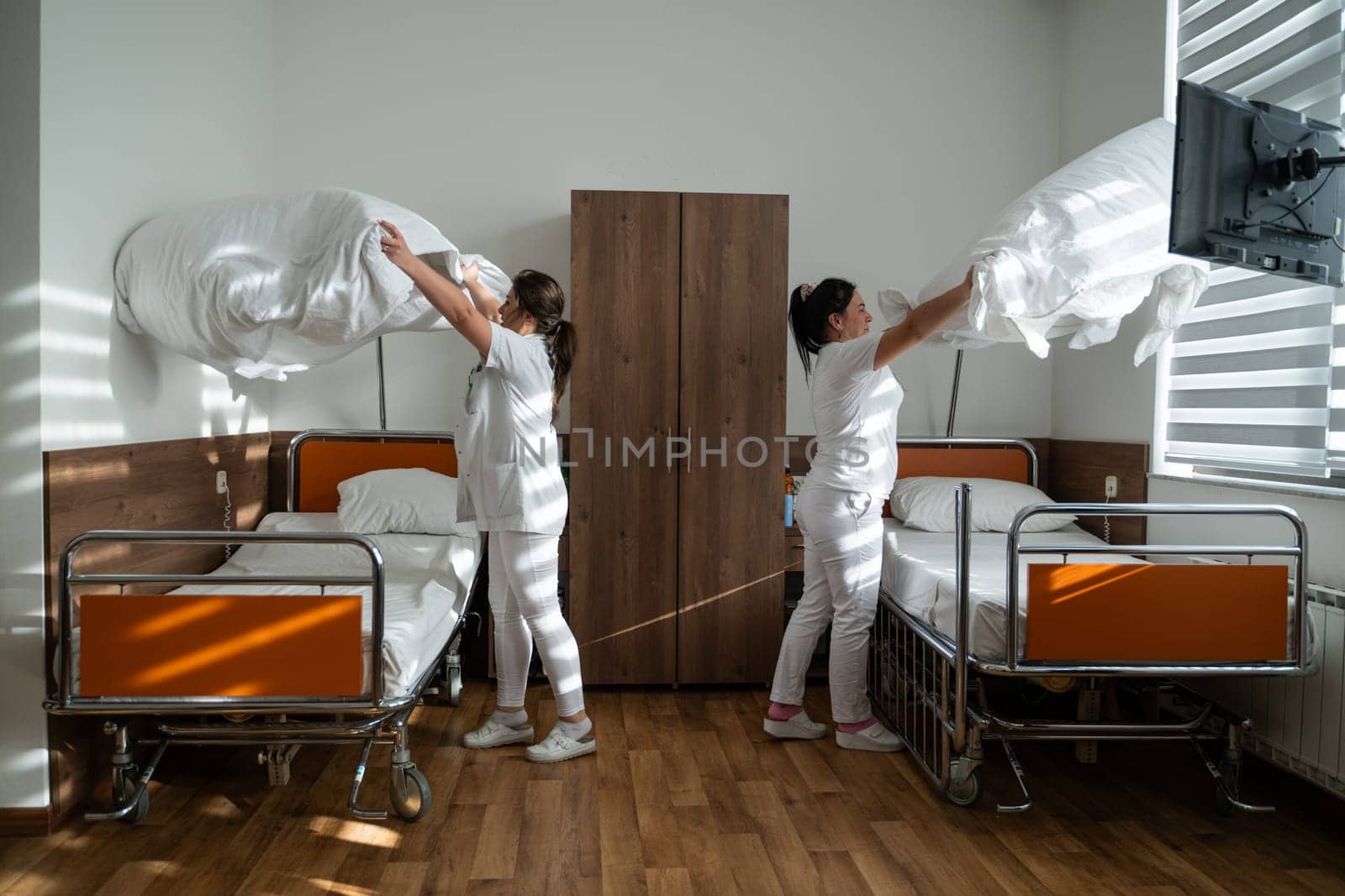 Two nurses in uniforms cleaning and organizing a hospital room by changing the bed linens and ensuring a hygienic environment for patients