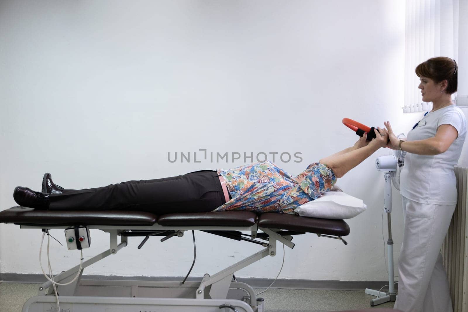 Elderly woman undergoing physiotherapy under the supervision of a female therapist. The patient is lying on a treatment table while performing an arm exercise with an orange pilates ring.