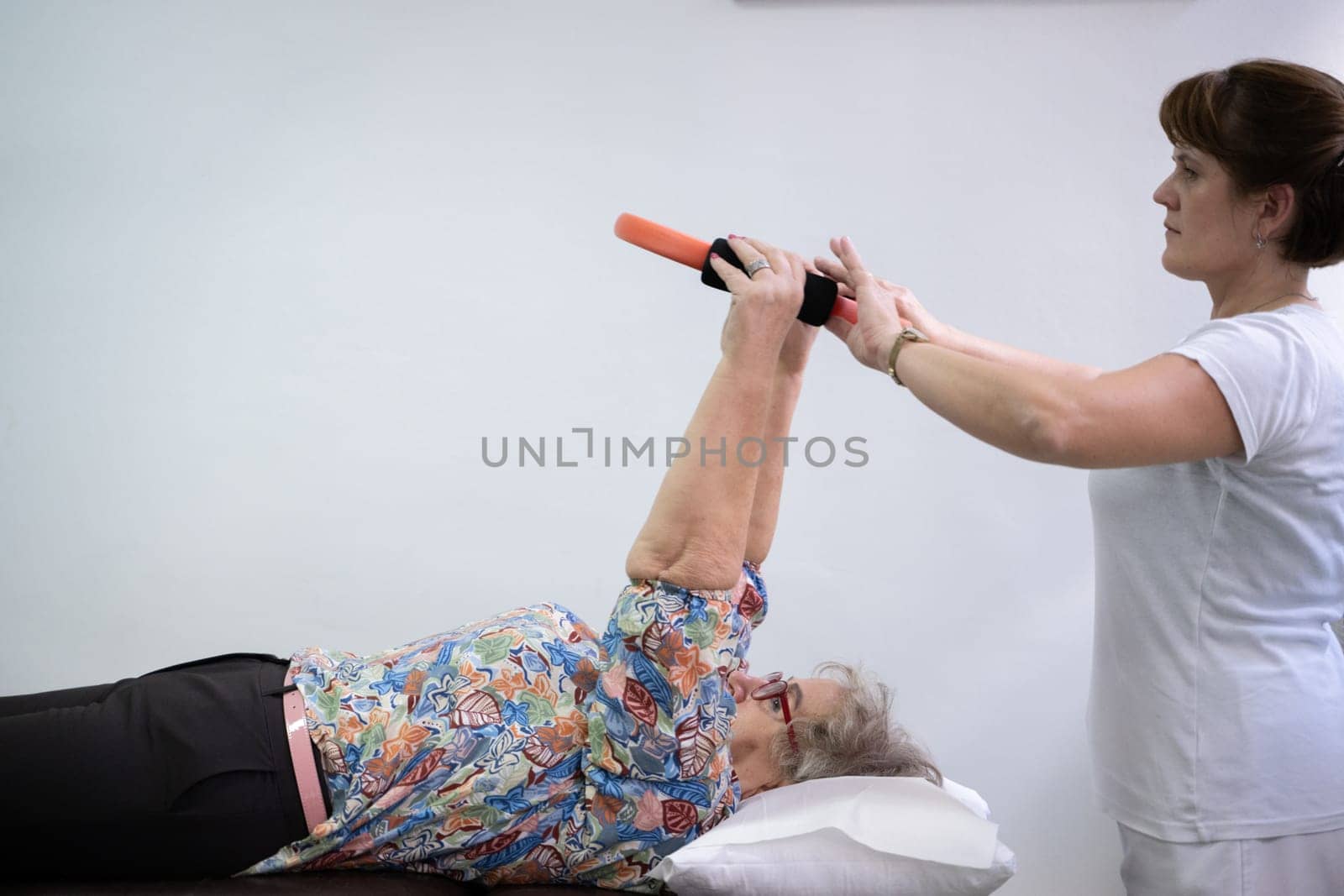 Elderly woman undergoing physiotherapy under the supervision of a female therapist. The patient is lying on a treatment table while performing an arm exercise with an orange pilates ring.