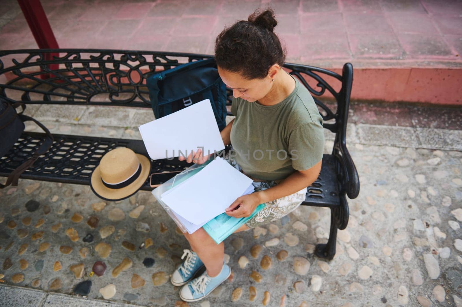 A woman sits on a decorative bench outdoors, examining documents. The scene conveys focus and tranquility, with accessories like a hat and backpack enhancing the casual ambiance.