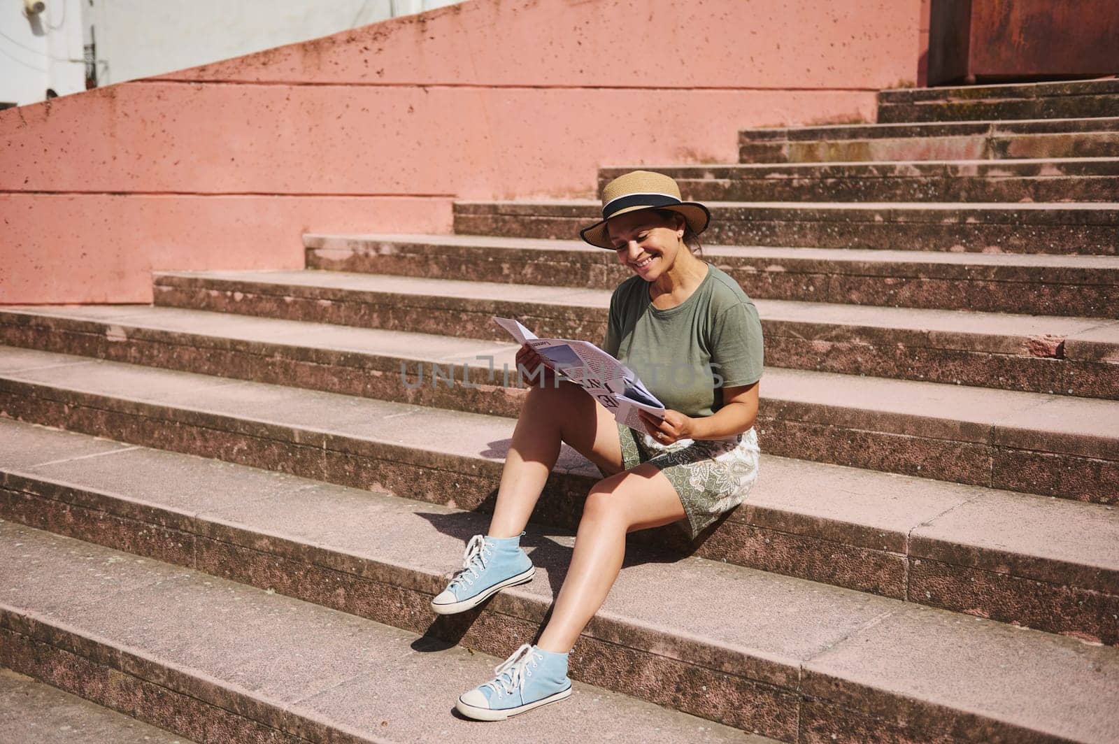 A cheerful woman sitting on outdoor steps, relaxing while reading a magazine and enjoying the sunny weather. Dressed casually, wearing a hat, this represents leisure, joy, and relaxation.
