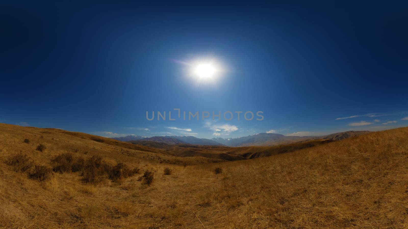 A wide panoramic view of arid, yellow-brown rolling hills under a clear blue sky with the sun shining.