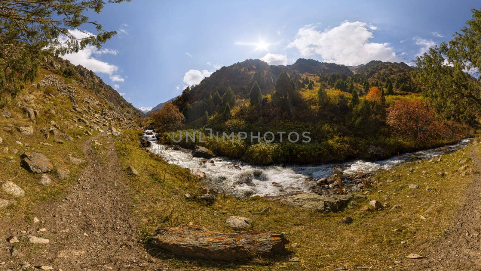 Low angle view upstream along a rushing river in Zhylamysh Gorge, Kyrgyzstan. Banks display vibrant autumn foliage and conifers below steep mountains.