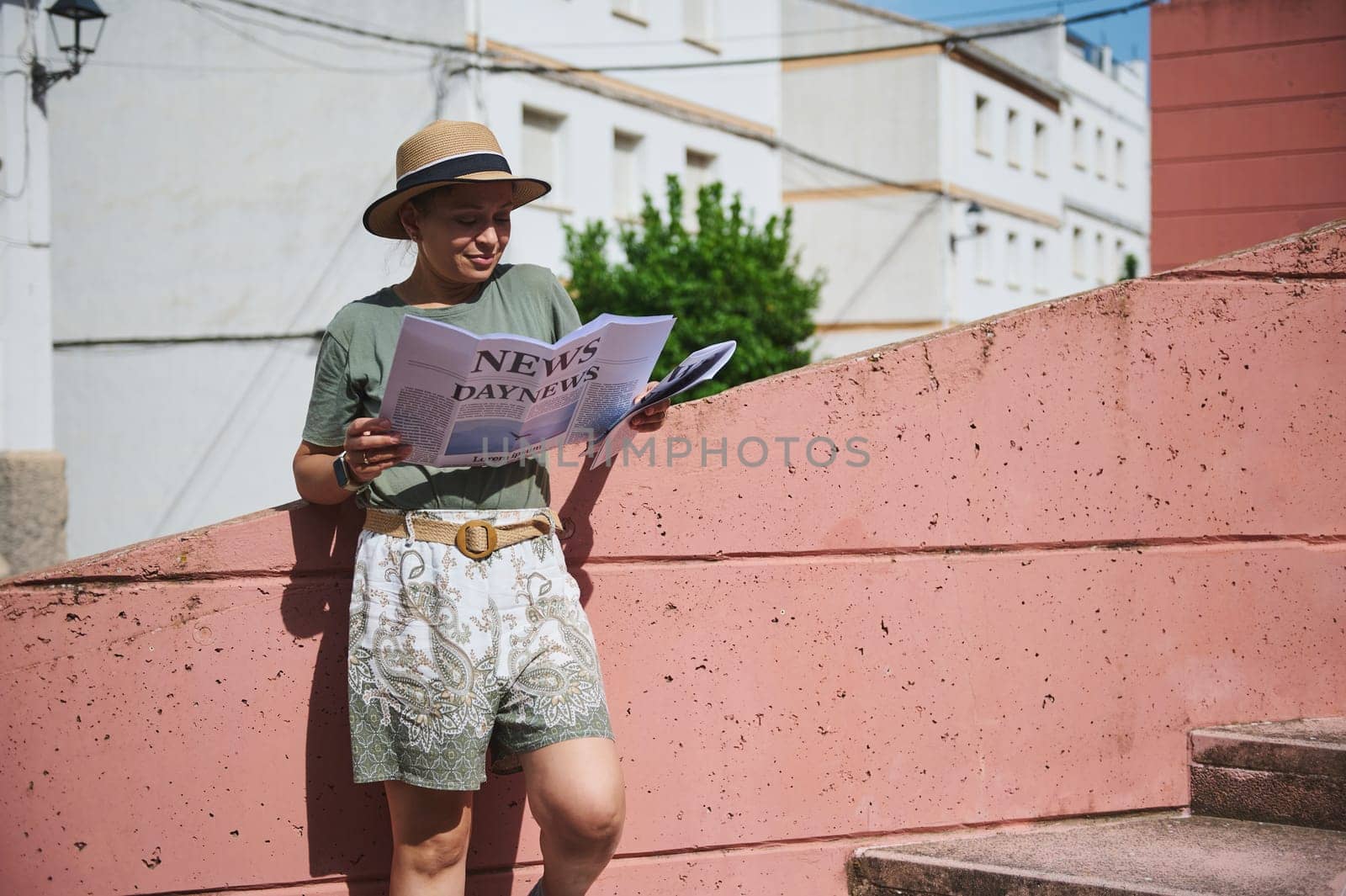 A casual moment outdoors as a woman in a hat and summer attire reads a newspaper against a bright wall under the sunlight, exuding relaxation and leisure.