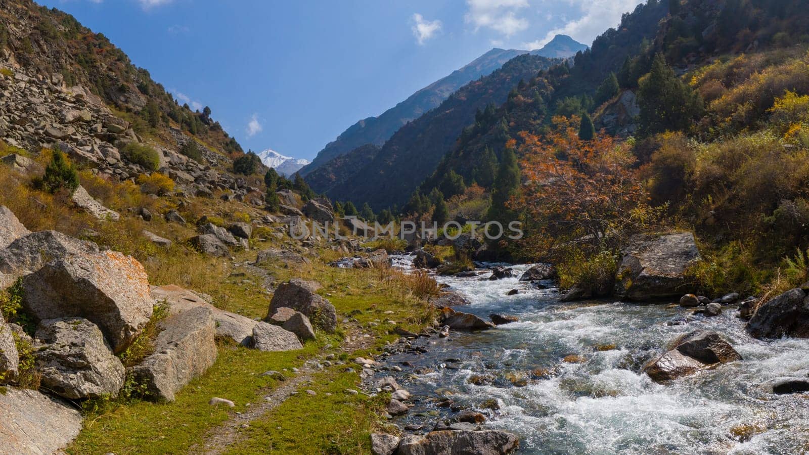 A wide shot captures a vibrant mountain river cascading over rocks, flanked by steep, rocky banks covered with patches of green and yellow vegetation.