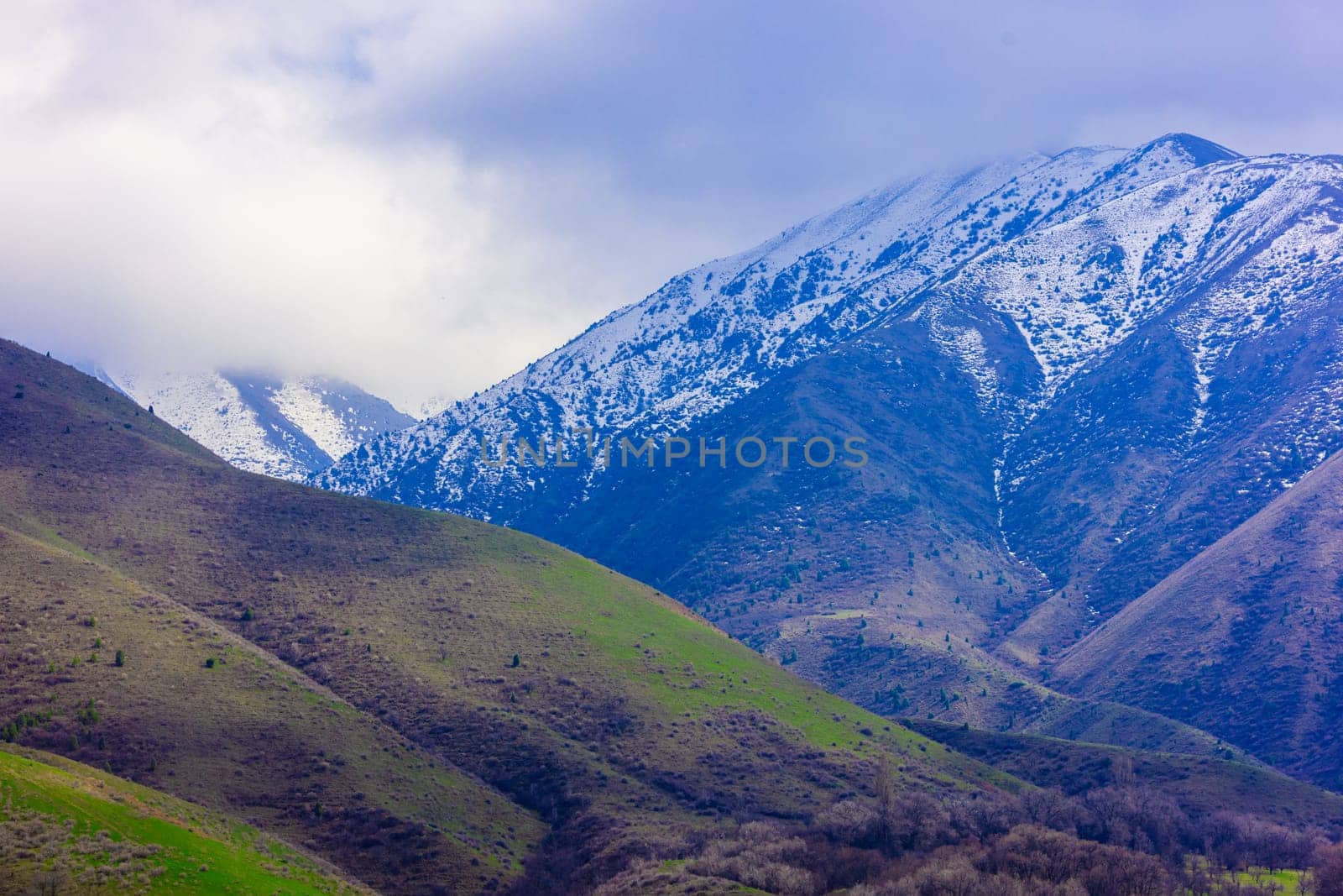 Snow-capped mountains in springtime Kyrgyzstan under a cloudy sky. Green and brown slopes.