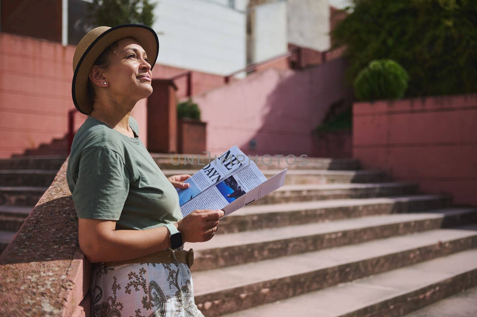 A cheerful woman in casual attire, enjoying a sunny day outdoors while reading a brochure. She exudes positivity as she leans against architectural stairs in a warm urban setting.