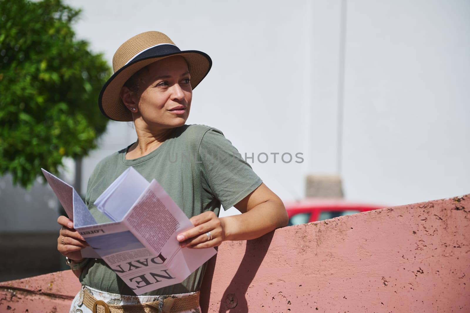 A woman in casual clothing and a straw hat holds a map while standing against a pink wall in a sunlit outdoor setting.