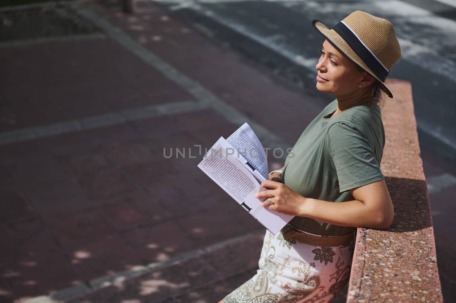 A content woman in a casual green shirt and straw hat enjoys reading a magazine while leaning against a wall on a sunny day outdoors.