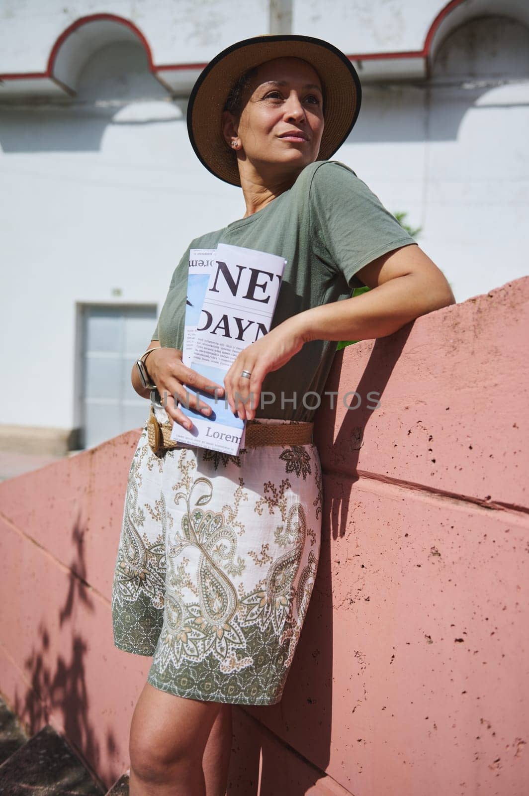 A fashionable woman wearing a straw hat and patterned shorts leans against a wall, holding a book, exuding relaxed confidence in the sunlit outdoor environment.