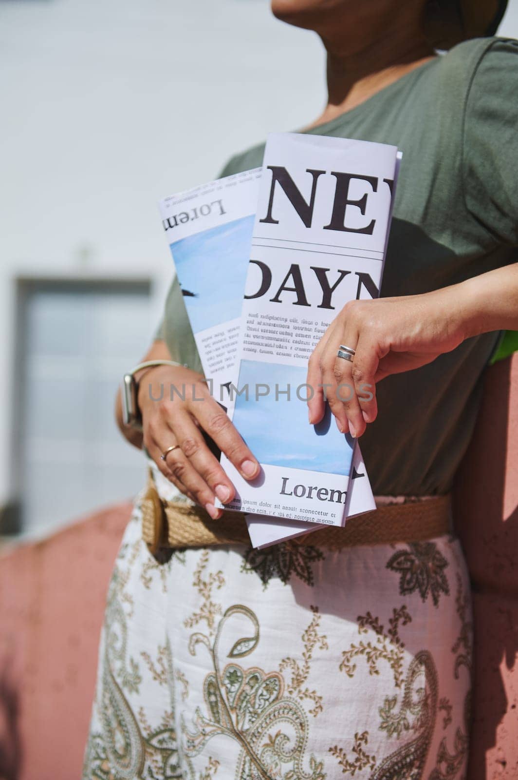 A stylishly dressed person holds a folded paper, featuring a printed text and design, while enjoying an outdoor setting in the sunlight.