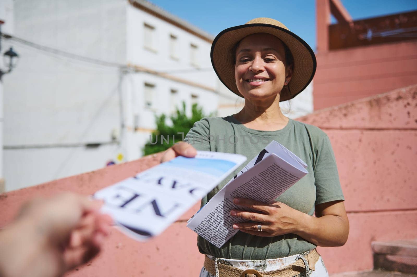 A cheerful woman wearing a hat and green shirt hands out flyers outdoors in a sunny urban setting. The image conveys friendliness, engagement, and community interaction.