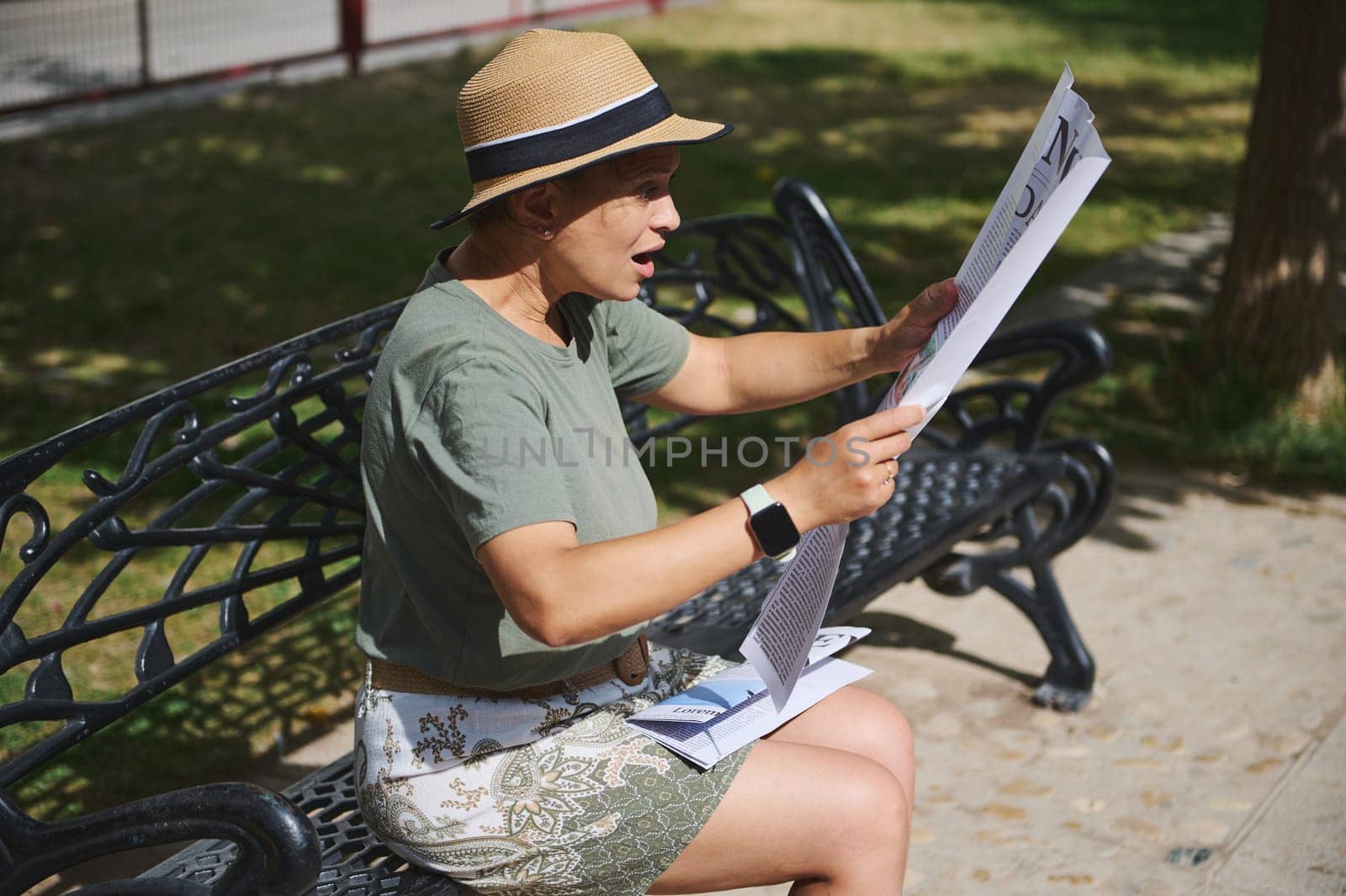 A woman enjoys reading a newspaper while sitting on a bench in a park. She appears engaged, wearing casual attire, under pleasant sunlight in a serene outdoor setting.