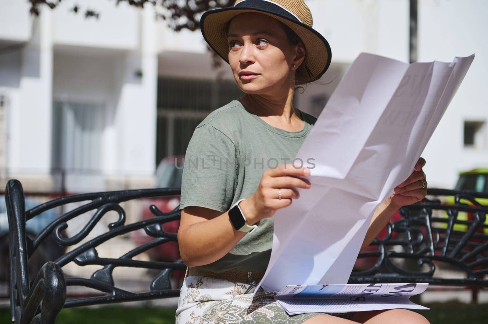 A thoughtful woman sits on a park bench while reading a large newspaper, enjoying the sunshine. She is wearing a straw hat and casual attire in a serene urban environment.