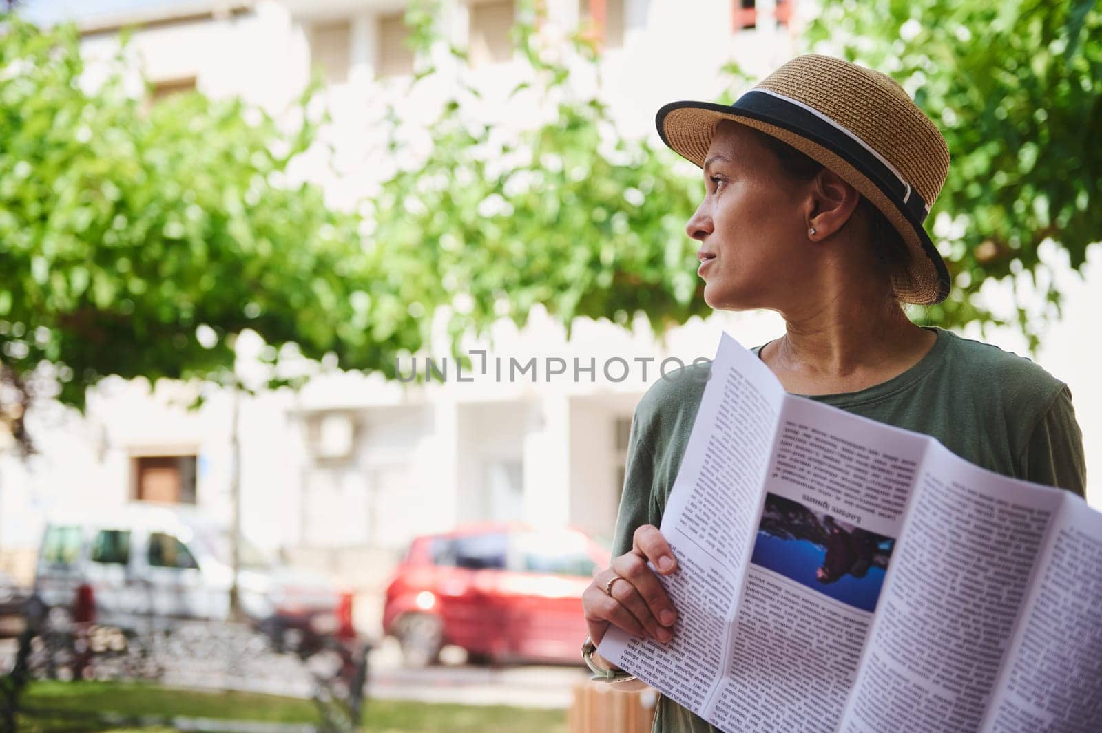 A woman wearing a hat with a magazine in hand, enjoying a quiet moment outdoors under the trees, surrounded by natural light and urban elements.