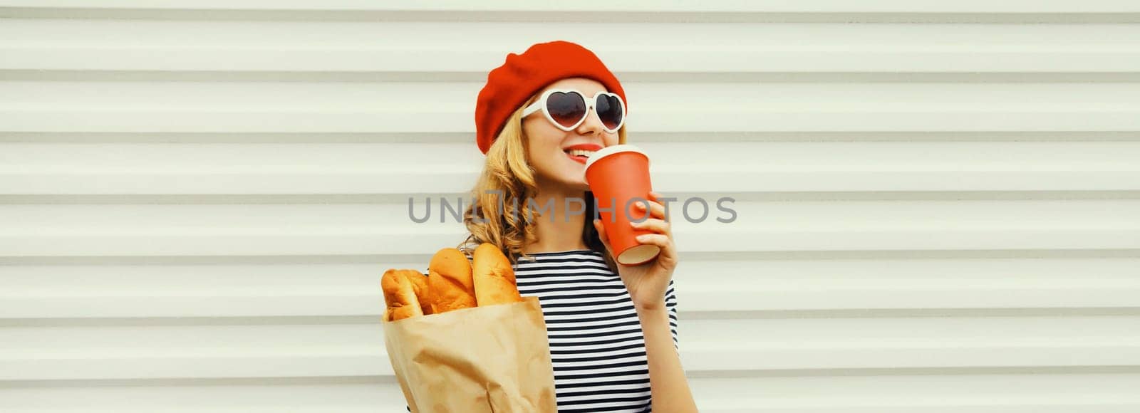 Beautiful happy smiling woman holding grocery shopping paper bag with fresh long white bread baguette, cup of coffee wear red french beret on white background