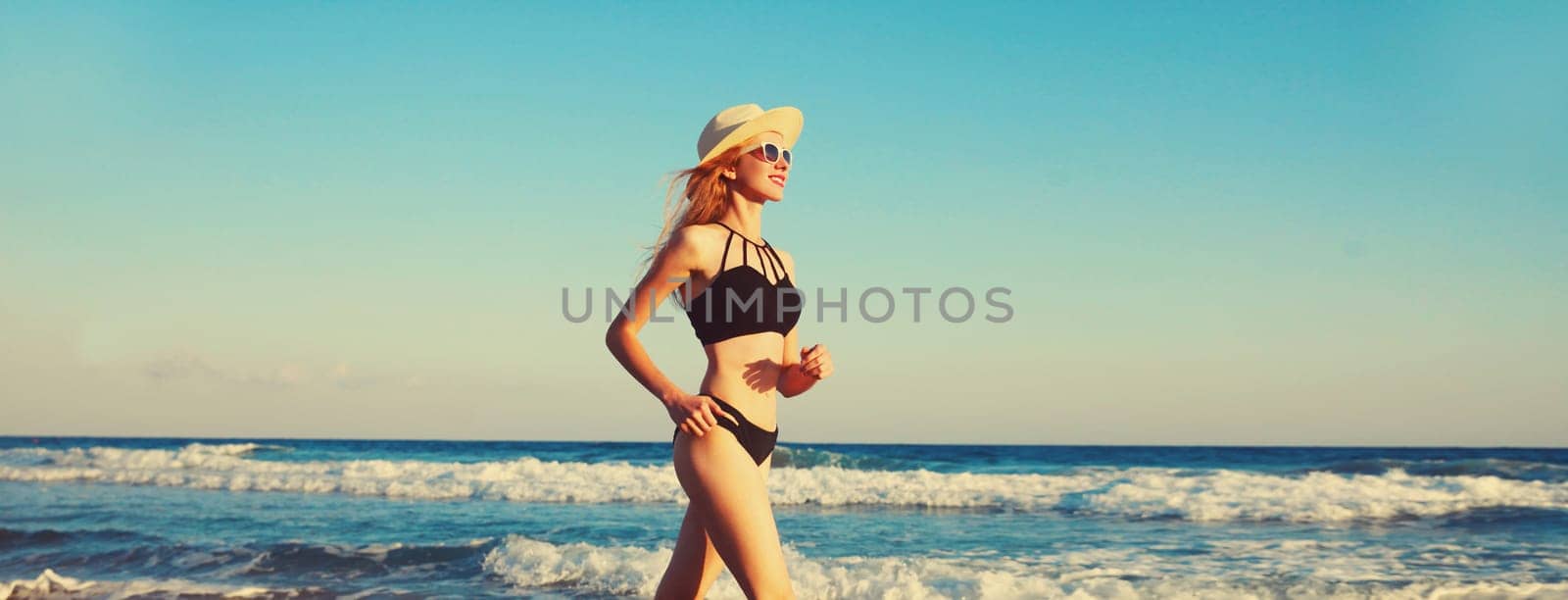 Summer vacation, active slim woman walking running in bikini swimsuit and tourist straw hat on the beach on sea coast with waves background on sunny day