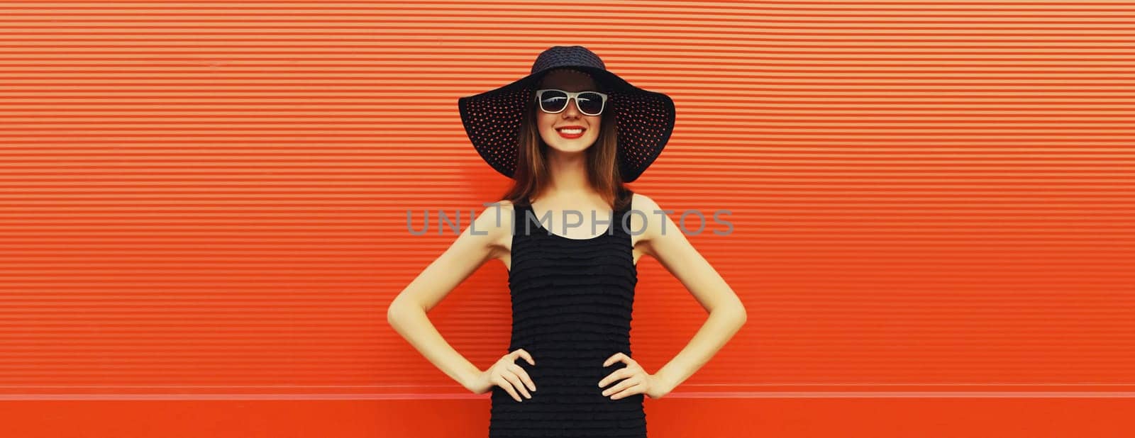 Summer portrait of beautiful stylish young woman posing in black straw hat, glasses, dress on bright red background