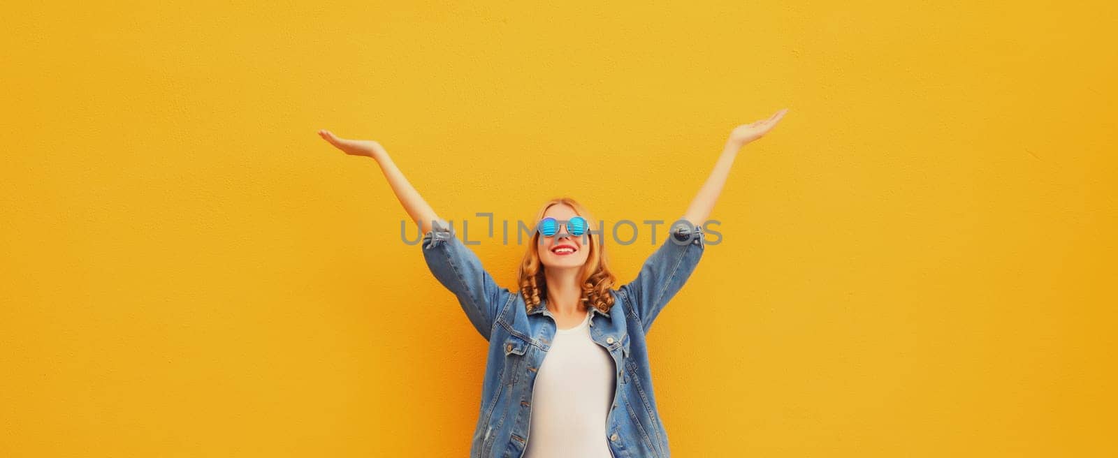 Summer holidays, inspired happy cheerful smiling young woman raising her hands up wearing denim clothing, glasses on colorful yellow background