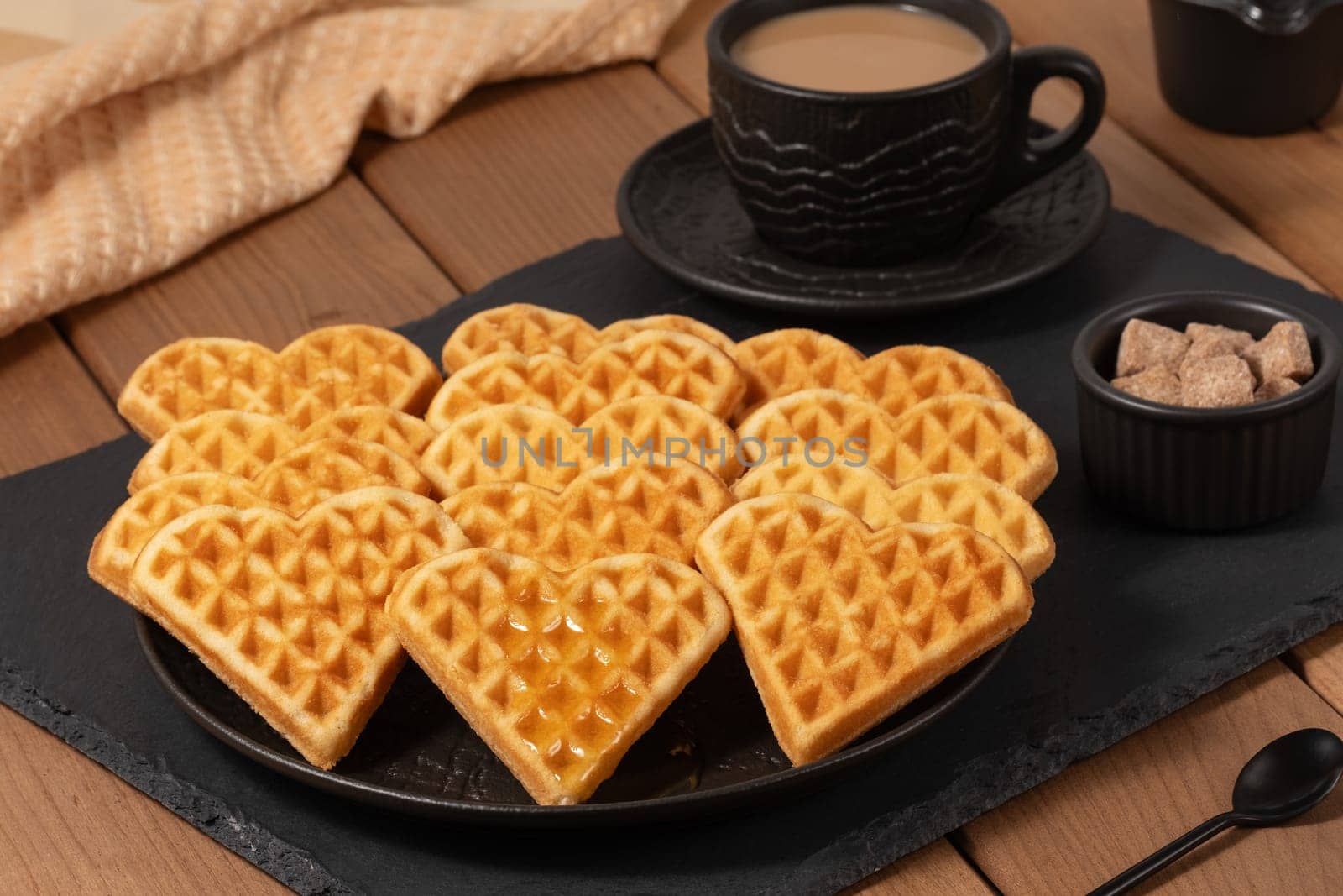Heart-shaped waffles are elegantly arranged on a black plate, accompanied by a cup of steaming coffee and a small bowl of sugar cubes, creating a warm and inviting atmosphere.