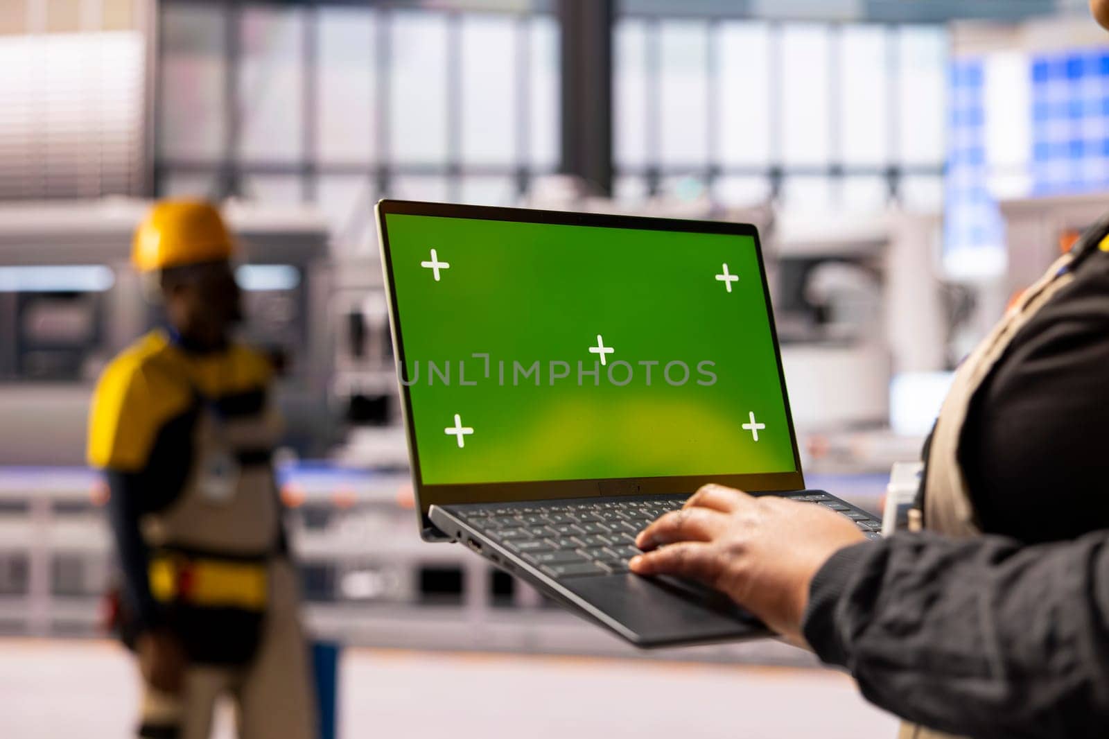African american factory worker holds laptop with green screen display, overseeing automated production line. The industrial plant integrates robotics, conveyor belts and smart manufacturing systems.