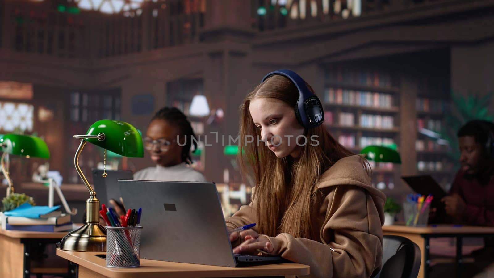 Young girl listening to educational documentary on laptop and takes notes, using modern non conventional way to study in the university library. Female student collects data. Camera A.
