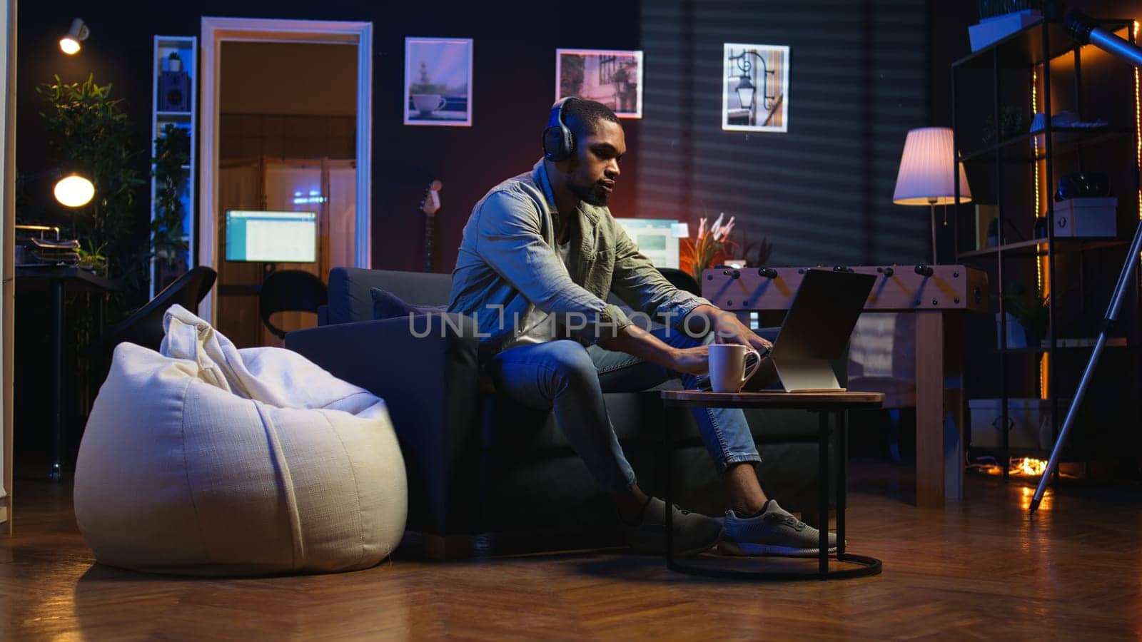 Male freelancer multitasking at home solving tasks with music on headset, listening to his favorite songs and paying attention to his remote work. African american guy enjoys schedule. Camera A.