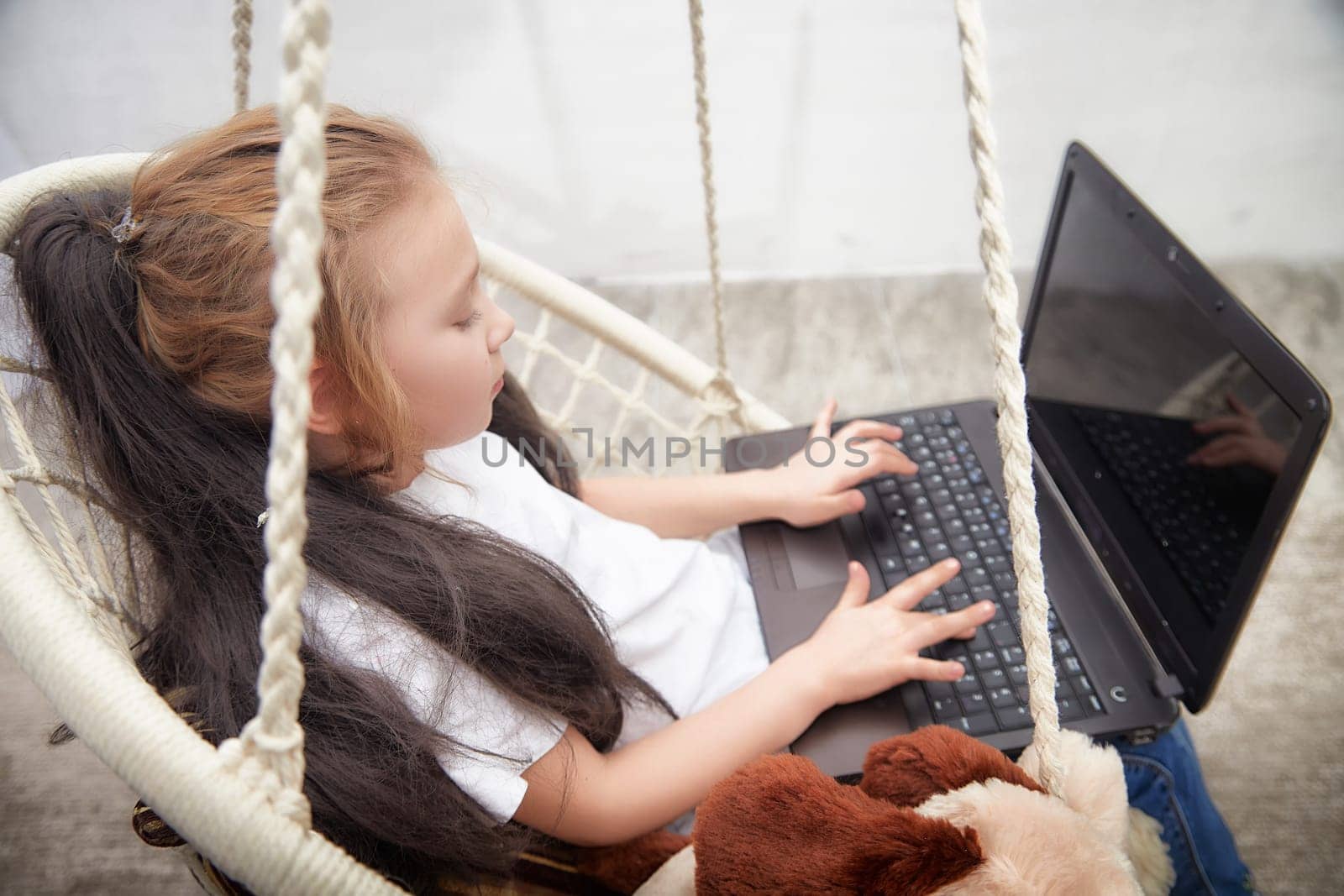 Young child happily types on a laptop while comfortably seated on a swing, engaged in a creative activity during afternoon hours.