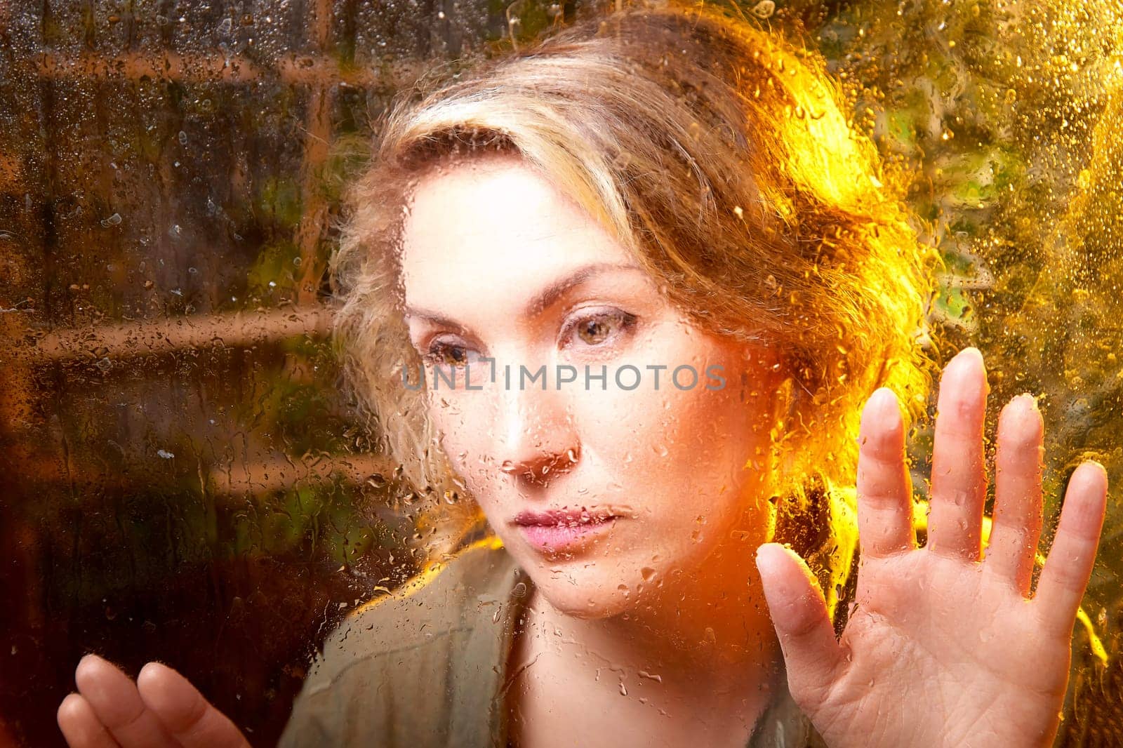 A woman presses her hands against a fogged window, lost in thought as warm light creates a comforting ambiance in the room.