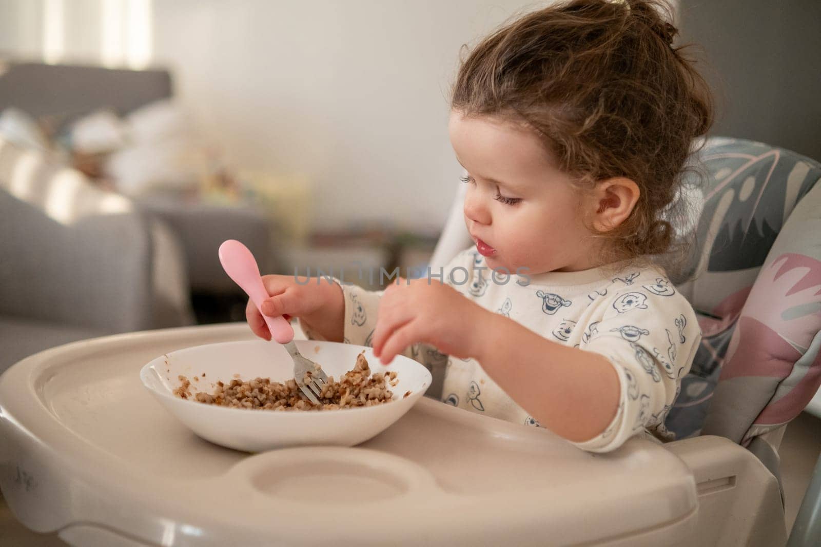 Happy toddler with curls having breakfast in a high chair. Kitchen background with bowl, spoon, and loving family environment.