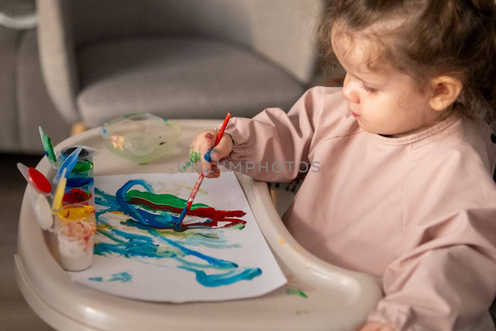 Little girl painting with gouache at a small childrens table. Bright room, toddler engaged in creative activity with colorful paints.